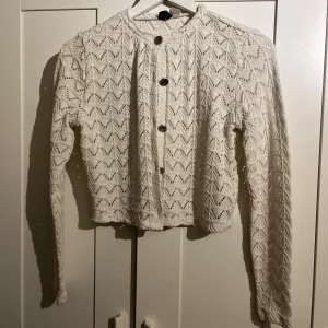 Vit stickad kofta med mönster - Supersöt vit stickad kofta med snyggt hålmönster och dekorativa knappar framtill. Koftan har lång ärm och rund halsringning, perfekt att slänga över en topp för en chill look. Mjuk och skön känsla, passar till både jeans och kjol.