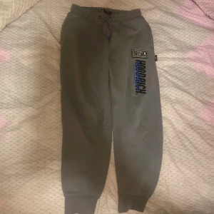 Grå mjukisbyxor från Hoodrich XS - Grå mjukisbyxor från Hoodrich i storlek XS med tryck och broderade detaljer i svart och blått. Byxorna har resår och snörning i midjan samt muddar vid bensluten. Perfekta för en chill och avslappnad stil.