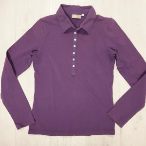 Långärmad tröja med krage - En lila långärmad tröja med krage och knappar framtill. Tillverkad i mjuk bomull (92 %) och elastisk elastan (8 %). Perfekt för dig som gillar stilrena plagg. Tröjan har naturligtvis tvättats. Se bilder för skick och passform. Storlek M enligt lappen men då måttet armhåla till armhåla är cirka 43 cm så stämmer S bättre. Axelsöm till ärmslut är cirka 60 cm. Tvättas i 60 grader.