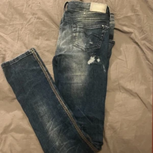  slitna jeans från Diesel - Säljer ett par blå jeans från Diesel med slitningar och hål på benen för en edgy look. Jeansen har normal passform, klassisk femficksdesign och dekorativa nitar längs sidsömmarna. Materialet är jeans i bomull och de har en snygg tvättad finish.