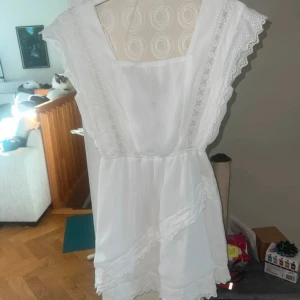 Vit kortklänning med spets och knyt - Superfin vit kortklänning med spetsdetaljer längs axlar och framstycke. Klänningen har omlottvolanger nertill och en knyt design i ryggen. Klänningen är av märket Dennis maglic i storlek S, fint skick och sparsamt använd 