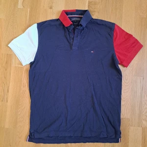 Tommy Hilfiger färgblockad piké M - Snygg pikétröja från Tommy Hilfiger i marinblått med röd och vit ärm samt tvåfärgad krage. Klassisk passform med knappar framtill och diskret logga på bröstet. Perfekt för dig som gillar sportig och stilren look.