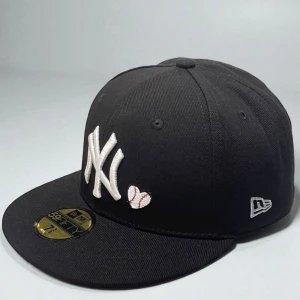 Svart New Era Yankees keps 59FIFTY 7 3/8 58.6 cm  - Säljer en svart New Era 59FIFTY keps med New York Yankees-logga i vitt framtill och ett broderat hjärta med baseball-sömmar. På sidan finns en färgglad World Series-patch och New Era-logga. Klassisk flat peak och stilren look, perfekt för dig som gillar streetwear.