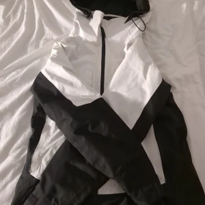 Svartvit vindjacka från Everest - Snygg svartvit vindjacka från Everest med huva och dragkedja framtill. Jackan har sportig design med svarta och vita paneler, samt praktiska fickor med dragkedja. Perfekt för dig som gillar stilren och funktionell ytterklädsel. Det är en skidjacka perfekt för vintern.