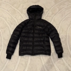 Svart Peak Performance Jacka - Säljer en svart dunjacka från Peak Performance med huva och dragkedja framtill. Jackan har quiltad design, ribbade muddar och två sidofickor med dragkedja. Perfekt för kalla dagar och har en clean look med broderad logga på bröstet. Använd 2,3 gånger. 170 men passar som S.(Ny pris: 3 199kr. Mitt pris: 699)