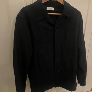 Marinblå  overshirt från Uniqlo - Stilren marinblå overshirt från Uniqlo med klassisk krage, bröstficka och knäppning framtill. Skjortan har lång ärm och en rak, lite loose passform. Perfekt att slänga över en t-shirt för en clean look.