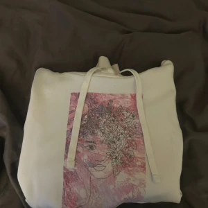 Vit hoodie med konsttryck och huva - Vit hoodie med stor ficka framtill och snörning vid huvan. På bröstet finns ett rosa konsttryck med ansikte och blommor, vilket ger en unik vibe. Mjuk och skön, perfekt för chill dagar. Liten svart etikett på ryggen.