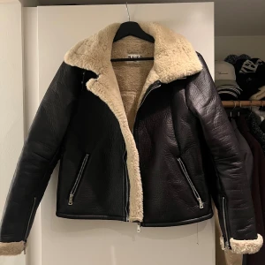 Svart pälsjacka från NA-KD - Säljer en svart pälsjacka från NA-KD med fluffig beige fuskpäls på krage och insida. Jackan har grov struktur, dragkedja framtill och på fickorna samt detaljer vid ärmslut. Perfekt för dig som vill ha en cool och varm look i vinter.