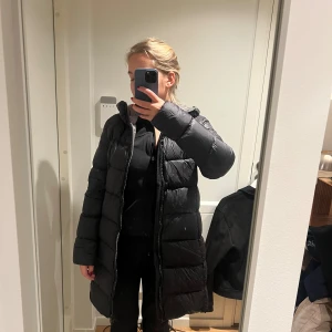 Svart lång dunjacka från Colmar - Säljer en svart, lång dunjacka från Colmar med huva och snyggt logomärke på ärmen. Jackan har dragkedja hela vägen och är quiltad för extra värme. Perfekt för kalla dagar och har en clean, stilren look som funkar till allt.