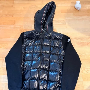 Svart Moncler cardigan med huva - Snygg svart cardigan från Moncler med glansig quiltad framsida, stickade ärmar och huva med röd och vit kant. Jackan har dragkedja framtill och Moncler-logga på ärmen. Perfekt för dig som vill ha en stilren och sportig look.
