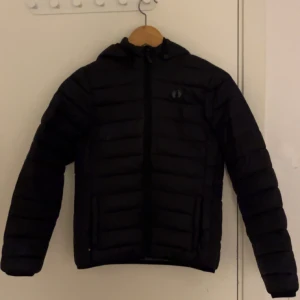 Svart pufferjacka från Hangten - Svart pufferjacka från Hangten med huva och horisontella quiltade paneler. Jackan har dragkedja framtill och två sidofickor med dragkedja. Perfekt för kalla dagar och har en clean, modern look som funkar till streetwear. Passar med allt.