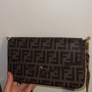 Fendi brun axelväska med FF-mönster - Snygg brun axelväska från Fendi med ikoniskt FF-mönster i svart. Väskan har en envelope-formad front med guldfärgad knapp och bred axelrem med matchande logga. Materialet är slitstark syntet och detaljerna är i guldmetall. Perfekt för dig som gillar statement-accessoarer.