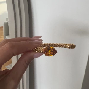 Guldarmband Caroline Svedbom med kristall - Unikt armband från Caroline Svedbom i guldton med pärlstruktur och en stor droppformad orange kristall. Armbandet är öppet och har märkets logga ingraverad på ena sidan. Perfekt statement piece för dig som vill sticka ut.