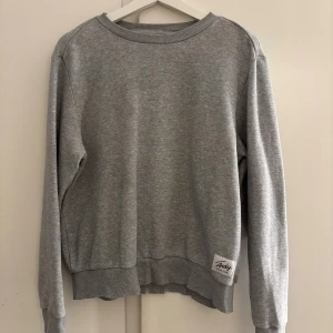 Grå sweatshirt  - En stilren grå sweatshirt från Andy by Frank Dandy i storlek S. Tröjan har rund hals, ribbade muddar och en snygg patch nertill. Perfekt för en chill och avslappnad look.🤗
