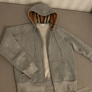 Grå Burberry hoodie med dragkedja - Säljer en grå hoodie från Burberry med klassiskt rutigt foder i huvan och broderad logga på bröstet. Tröjan har snörning i huvan, två fickor fram och silverfärgad dragkedja med Burberry-gravyr. Mjuk insida och ribbade muddar för extra comfort.
