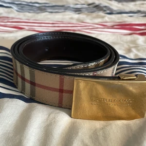 Burberry rutigt bälte med guldspänne - Snyggt bälte från Burberry med klassiskt rutig design i beige, rött och svart. Bältet har ett rejält guldigt spänne med Burberry-logga ingraverad. Tillverkat i skinn och levereras med originalpåse och box. Perfekt accessoar för att lyfta din outfit. Pris kan förhandlas 