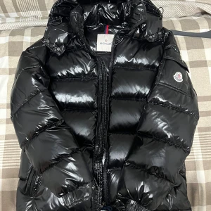 Svart Moncler pufferjacka med huva - Säljer en svart pufferjacka från Moncler Storlek S med glansig finish och huva. Jackan har dragkedja framtill, ficka med Moncler-logga på ärmen och quiltad design. Perfekt för dig som vill ha en snygg och varm jacka med street vibe.