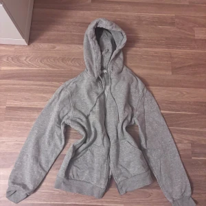 Grå hoodie från H&M med dragkedja - Grå hoodie från H&M med huva och snörning, hel dragkedja framtill och två fickor. Tröjan har ribbade muddar vid ärmslut och nederkant, samt långärmad passform. Perfekt för chill dagar och enkel att matcha med jeans eller joggers. (Saknar dragkedja och har en målarfläck)