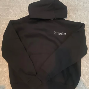 NY Svart hoodie Dropsize  - Svart hoodie från Dropsize med tryck på bröstet och stor grafik på ryggen med text, duvor och coola detaljer. Tröjan har huva, lång ärm och en klassisk känguruficka framtill. Perfekt för dig som gillar streetwear och statement-plagg.