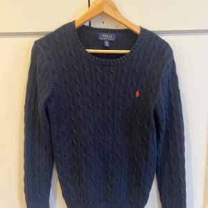 Marinblå stickad tröja Polo Ralph Lauren - Snygg marinblå stickad tröja från Polo Ralph Lauren med klassiskt kabelstickat mönster och rund hals. Tröjan har långa ärmar och den ikoniska röda logon broderad på bröstet. Perfekt för dig som gillar stilrena och tidlösa plagg.