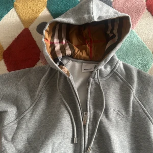 Grå hoodie från Burberry med dragkedja - Säljer en grå hoodie från Burberry i storlek M. Tröjan har klassisk Burberry-rutig huva i brunt, beige, svart och rött, snörning och dragkedja framtill. Mjuk bomullskänsla och broderad logga på bröstet. Perfekt för dig som gillar stilrena streetwear-vibes.