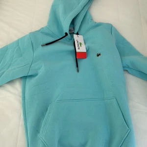 Ljusblå hoodie från Lacoste S - Snygg ljusblå hoodie från Lacoste i storlek S. Tröjan har huva med svarta snören, klassisk känguruficka och den ikoniska krokodilloggan på bröstet. Mjuk bomullskvalitet som är skön att ha på sig. Perfekt för chill dagar. 