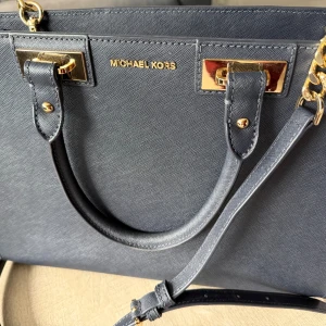 Blå handväska från Michael Kors - Stilren svart handväska från Michael Kors med guldfärgade metalldetaljer och kedja. Väskan har både handtag och justerbar axelrem, tillverkad i skinn med en elegant, strukturerad yta. Perfekt för dig som gillar klassisk design med lyxig touch. 33/24 cm