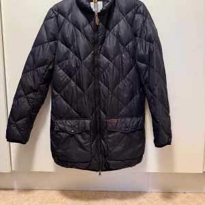 Peak Performance tunn dunjacka L - Tunn dunjacka från Peak Performance i storlek L. Style Joyce LO J. Mycket fint skick, väl omhändertagen. Jackan har snyggt rutmönster, två fickor med lock och knappar framtill samt dragkedja med läderdetalj. Materialet är 100% polyamid. 