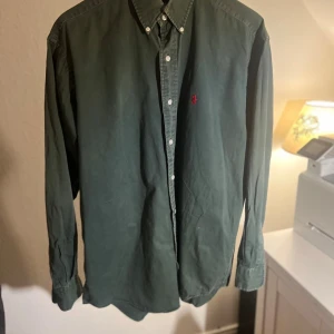 Grön skjorta från Ralph Lauren M - Klassisk grön skjorta från Ralph Lauren i modellen Blaire. Skjortan har knappar hela vägen, button down-krage och den ikoniska röda broderade loggan på bröstet. Tillverkad i 100% bomull och har långa ärmar. Perfekt för en stilren och avslappnad look.