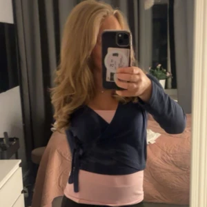 Mörkblå omlottopp med knyt - Säljer en mörkblå omlottopp med långa ärmar och knyt i sidan. Toppen är croppad och har en snygg v-ringad design som ger en cool och modern look. Perfekt att matcha med en ljusrosa topp under för extra kontrast.