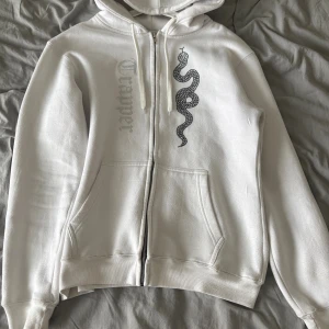Made by trappers vit hoodie - Vit hoodie från Trapper i storlek XL med dragkedja, huva och snörning. På framsidan finns ett svart ormtryck och texten 'Trapper' i silvriga paljetter. Tröjan har två fickor och är tillverkad i mjuk bomullsmix, perfekt för chill dagar.