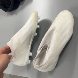 Vita adidas Crazyfast fotbollsskor - Säljer ett par vita adidas Crazyfast fotbollsskor med klassiska tre ränder på sidan. Skorna har en strumpliknande ovandel utan snörning och är tillverkade i syntetmaterial. Perfekta för dig som vill ha lättviktsskor med bra grepp på planen.