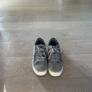 Grå sneakers med dragkedja och snörning - Säljer ett par grå sneakers från VOI med snygga dragkedjedetaljer på sidorna och klassisk snörning. Skorna har en vit platt sula och är gjorda i mocka-liknande material. Perfekta för dig som gillar en stilren och urban look.