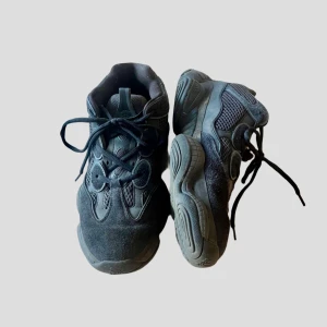 Yeezy 500  - Säljer ett par svarta Adidas Yeezy 500 sneakers med chunky sula och cool mesh- och mockadetaljer. Skorna har snörning och en futuristisk vibe som passar perfekt till streetwear. Grymt sköna och riktigt snygga på foten.