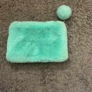 Turkos fluffig kuvertväska med boll - Supermjuk och fluffig turkos kuvertväska med dragkedja och en söt matchande boll som detalj. Perfekt storlek för det viktigaste och ger en lekfull touch till din outfit. Insidan är fodrad i grått tyg och väskan har en rektangulär form.