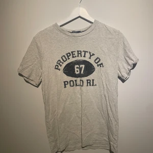 Grå t-shirt från Polo Ralph Lauren - Snygg grå t-shirt från Polo Ralph Lauren med tryck 'Property of 67 Polo RL' i svart på bröstet. Klassisk rund hals och korta ärmar. Tillverkad i 100% bomull. 