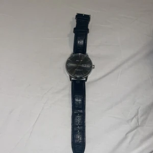  Silver klocka från GANT med svart läderband.   Äkta - Stilren klocka från GANT med rund silverfärgad urtavla och dag/datumvisning. Bandet är svart och har en snygg krokodilpräglad yta i äkta läder. Baksidan är i rostfritt stål och spännet är också märkt med GANT. Perfekt accessoar för dig som gillar klassisk design.