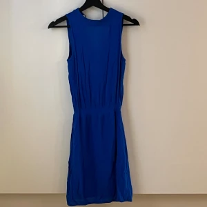 Blå ärmlös kortklänning - En stilren blå klänning med ärmlös design och rund halsringning. Klänningen har en markerad midja och lätt rynkade detaljer som ger en snygg siluett. Perfekt för dig som gillar enkel men snygg stil.
