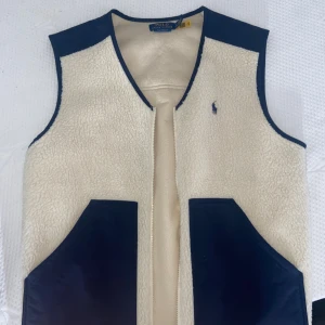 Beige fleeceväst från Polo Ralph Lauren - Snygg beige fleeceväst från Polo Ralph Lauren med marinblå detaljer och fickor. Västen har v-ringad hals och ett litet broderat Polo-logo på bröstet. Perfekt för lager-på-lager och chill stil under höst och vinter.