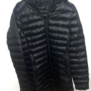 Svart dunjacka med huva - Svart dunjacka från Everest med huva och dragkedja framtill. Jackan har ett stilrent, quiltat mönster och är lätt vadderad för extra värme. Passformen är normal och materialet är syntet, vilket gör den både lätt och smidig. Perfekt för kyliga dagar. Nypris 999kr