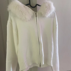 Vit hoodie med fuskpäls och dragkedja - Mysig vit hoodie med fluffig fuskpäls runt huvan och hel dragkedja framtill. Stickad känsla med ribbade muddar vid ärmslut och nederkant. Perfekt för dig som gillar en soft och stylish look under kyliga dagar.