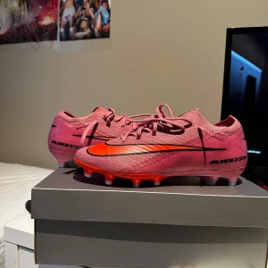 Nike Air Zoom rosa fotbollsskor - Säljer ett par Nike Air Zoom fotbollsskor i en snygg rosa färg med röda och svarta detaljer. Skorna har snörning, platt sula med dobbar och en sportig design. Ovandelen är i syntetmaterial för lätt känsla och bra passform. Perfekta för dig som vill sticka ut på planen.