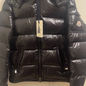 Svart pufferjacka från Moncler - Säljer en svart pufferjacka från Moncler med glansig finish och klassisk logga på ärmen. Jackan har dragkedja framtill, två fickor med dragkedja och quiltad design. Perfekt för dig som vill ha en snygg och varm jacka med street vibe. Helt ny oanvänd priset kan diskuteras vid snabb affär.