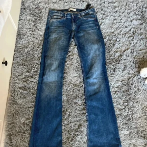 Blå bootcut jeans  - Snygga blå jeans med klassisk bootcut-passform. Jeansen har en lågmidjad midja, fem fickor och slitningar framtill för en cool, avslappnad look. Perfekta för dig som gillar en tidlös stil med lite extra edge.