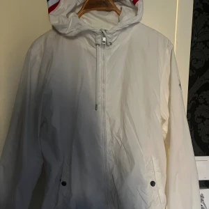 Vit vindjacka från Moncler med huva - Snygg vit vindjacka från Moncler med huva som har coola röd, vit och marinblå ränder. Jackan har dragkedja framtill, snörning vid halsen och Moncler-logga på ärmen. Perfekt för dig som vill ha en clean och sportig look.
