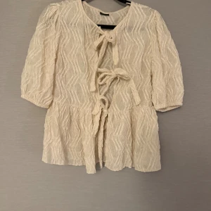 Krämvit blus med knyt - Supersöt krämvit blus med strukturmönster och tre knyt framtill. Blusen har trekvartsärmar och volang nedtill, vilket ger en luftig och romantisk vibe.