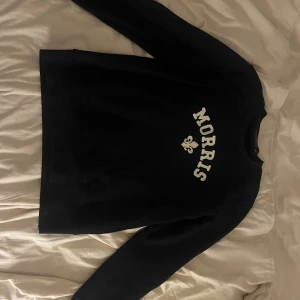Mörkblå  sweatshirt från Morris - Mörkblå sweatshirt från Morris med vit broderad logga och en fleur-de-lis på bröstet. Tröjan har rund hals och långa ärmar, perfekt för en chill och stilren look. Mycket bra skick. 
