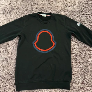 Svart sweatshirt från Moncler med logga - Svart sweatshirt från Moncler i bomull med stor broderad Moncler-logga i rött och blått på bröstet. Klassisk rund hals och långärmad modell. Moncler-märke på vänster ärm. Perfekt för dig som gillar stilrena designer med tydlig branding.