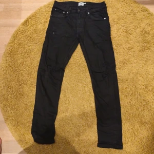 Svarta slim jeans från 157 - Svarta jeans från 157 med klassisk femficksdesign och smal passform. Jeansen är tillverkade i ett stretchigt bomullsmaterial som sitter tajt längs benen. Perfekta för dig som gillar en clean och enkel look. Har en defekt vid knäna där det fanns ett hål som nu är sydd