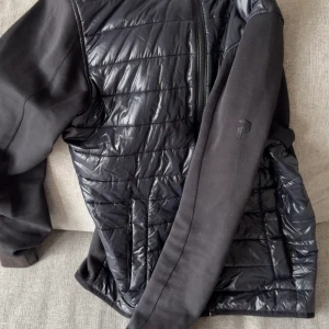 Svart hybrid jacka Peak Performance S - Svart hybrid jacka från Peak Performance i storlek S. Jackan har quiltad framsida med glansigt material och släta svarta ärmar och rygg. Dragkedja framtill och två fickor. Snygg, sportig look med diskret logga på ärmen.